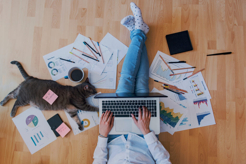 Woman working from home, sitting on the floor surrounded by papers, holding a laptop, with a cat resting near her.
