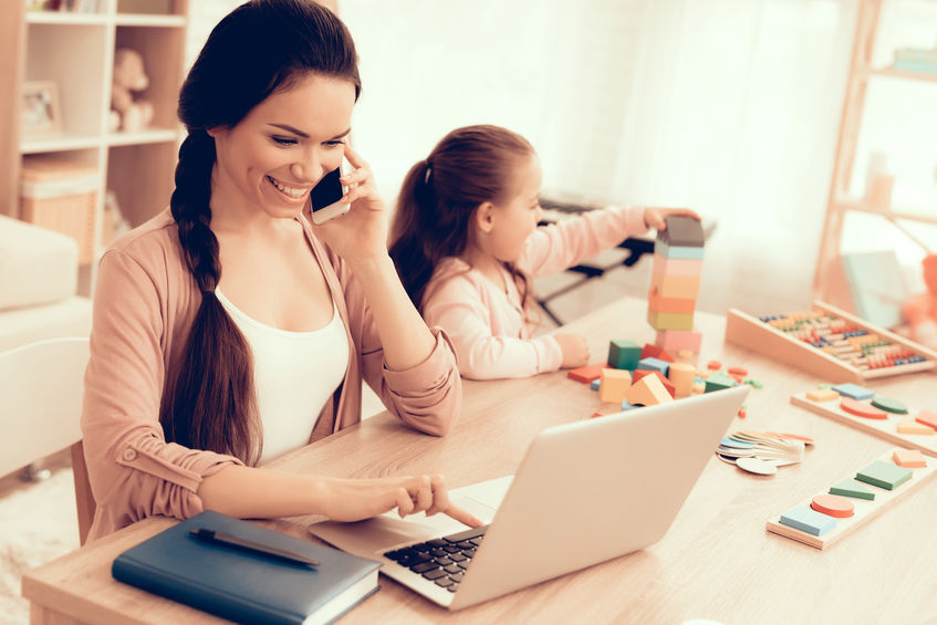 Mother at the table, on the phone and the laptop, working at home, while her daughter is playing an educational game next to her.